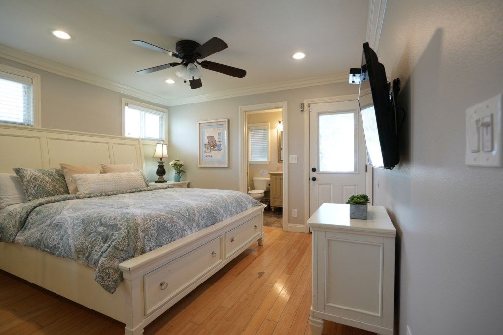 Un dormitorio con cama, ventilador de techo, televisor montado en la pared y puerta que da a otra habitación.