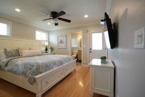A bedroom with a bed, a ceiling fan, a TV mounted on the wall, and a door leading to another room.
