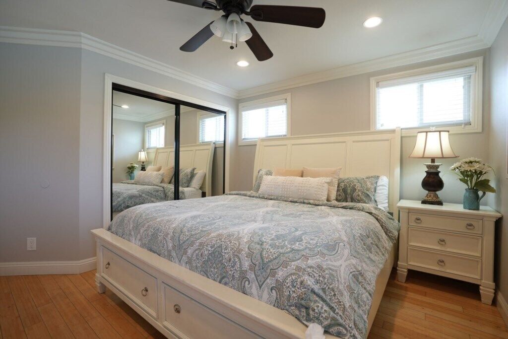 Une chambre à coucher avec un lit, des tables de chevet, un miroir, un ventilateur de plafond et une fenêtre.