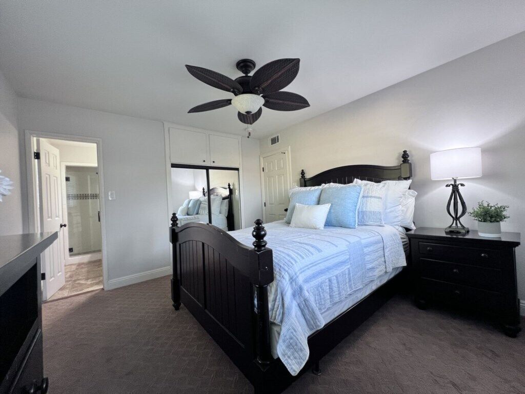 A bedroom with a bed, a ceiling fan, a bedside table with a lamp, and a door leading to another room.