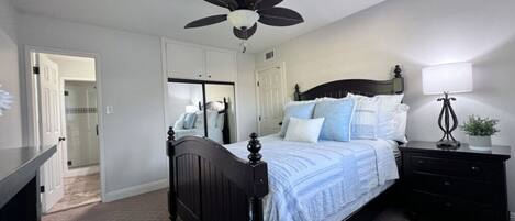 A bedroom with a bed, a ceiling fan, a bedside table with a lamp, and a door leading to another room.