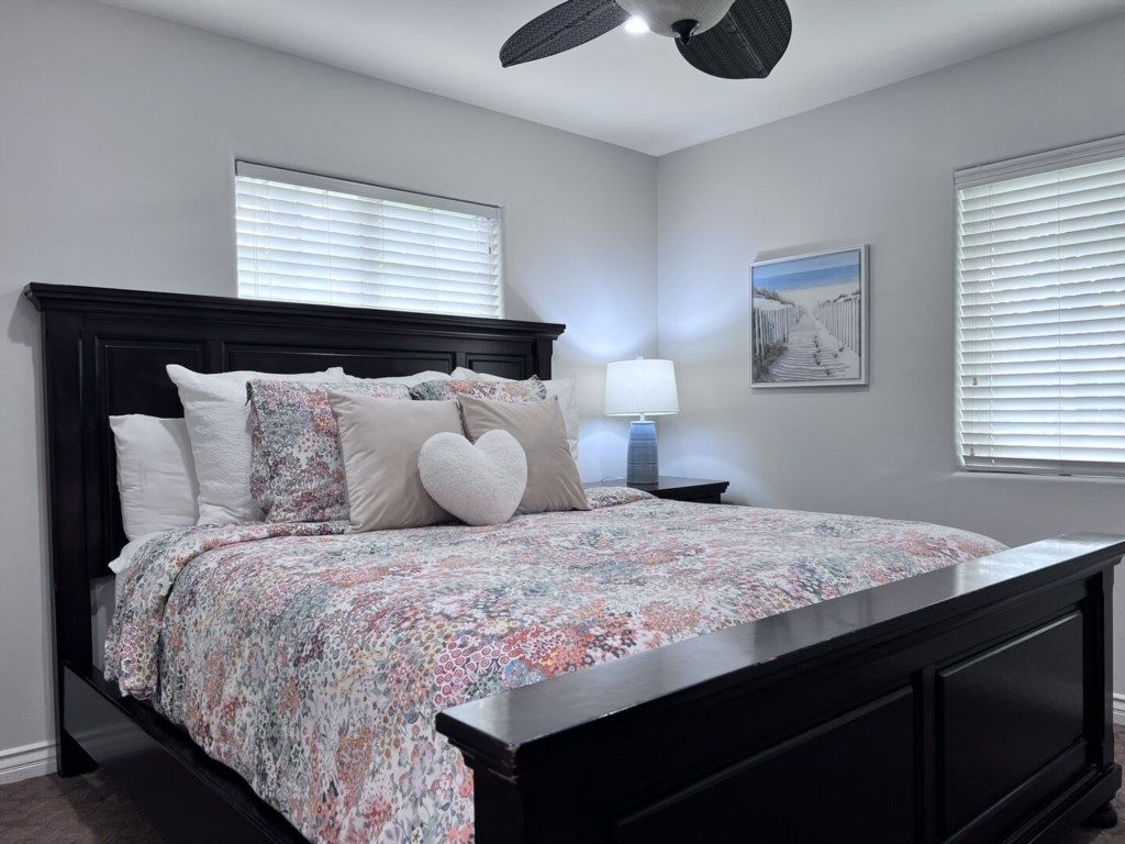 A bedroom with a dark wood bed, a floral bedspread, a nightstand with a lamp, and a framed picture on the wall.