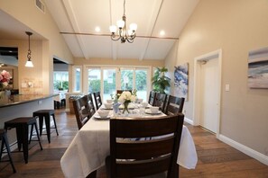 A dining area with a large table, chairs, and a chandelier. There is a kitchen area with a counter and stools. The room has large windows and a door. - Snazzy 6 Bedroom 3 Bath Pool Home 1/2 Mile Walk to Disney (Anaheim)