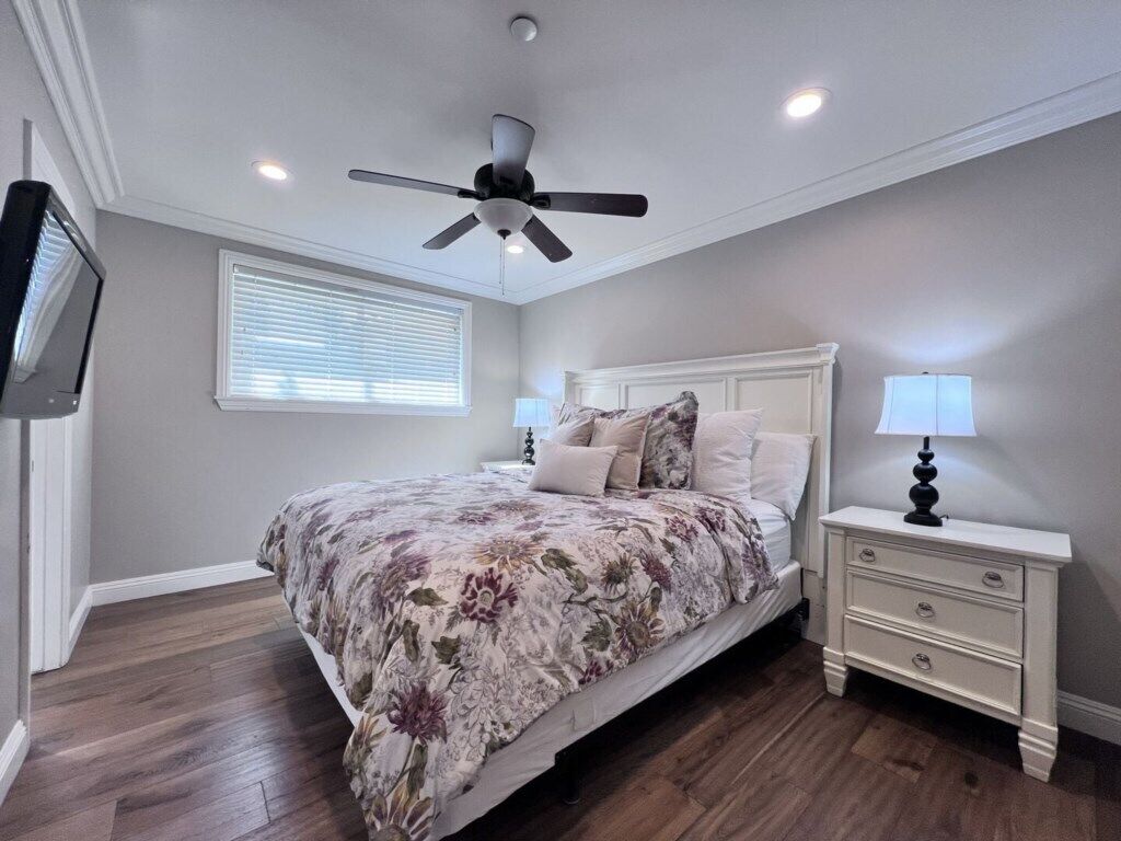 A bedroom with a bed, nightstands, a ceiling fan, a TV, and a window with blinds.