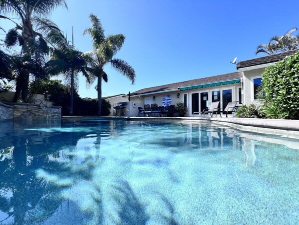 A clear swimming pool with a stone wall, palm trees, and a building with a tiled roof and outdoor seating.