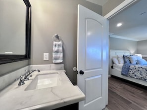 A bathroom with a white sink, a towel rack, and a door leading to a bedroom with a bed and pillows.