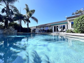 A clear swimming pool with a stone wall, palm trees, and a building with a tiled roof and outdoor seating.