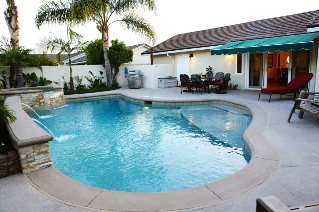 A swimming pool with a curved edge, surrounded by a patio area with chairs and a table.