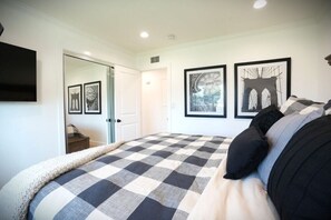 A bedroom with a checkered bedspread, framed pictures on the wall, a door leading to another room, and a flat-screen TV mounted on the wall.