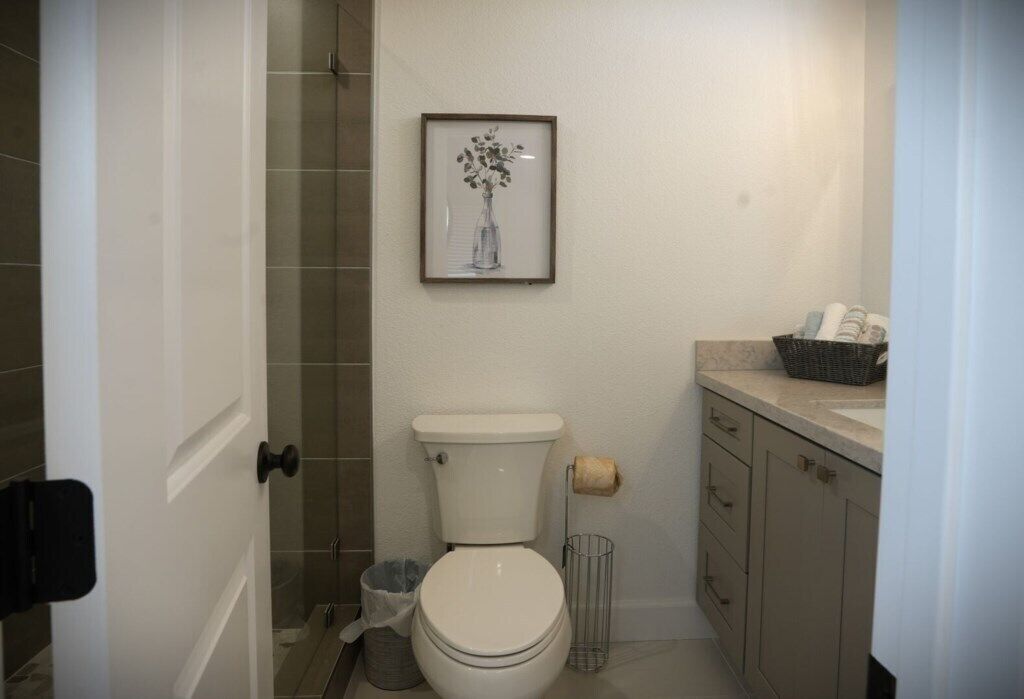 A bathroom with a toilet, a framed picture on the wall, a shower area, and a sink with a basket of toiletries.