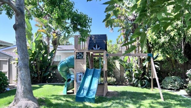 Ein Garten mit einer hölzernen Spielanlage, darunter eine Rutsche und ein Kletterbereich, umgeben von Bäumen und Rasen.