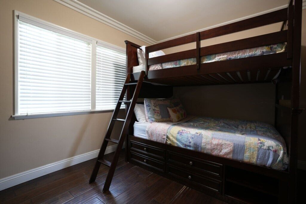 Una cama litera con escalera, una ventana con persianas y piso de madera.