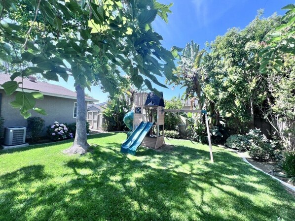 A backyard with a wooden play structure, a slide, and a house in the background. - Charming Anaheim Home 1/2 Mile Walk to Disney (Anaheim)