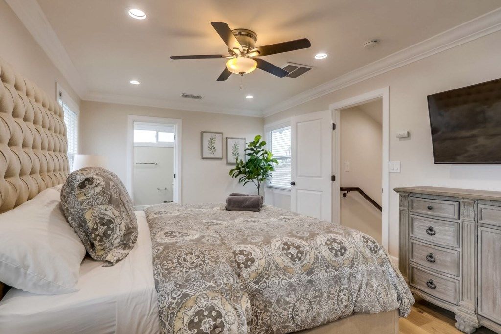 A bedroom with a tufted headboard, a patterned bedspread, a ceiling fan, and a dresser.