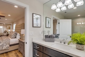 Un dormitorio con cama, mesitas de noche, cómoda, espejo grande y baño con lavabo y espejo.