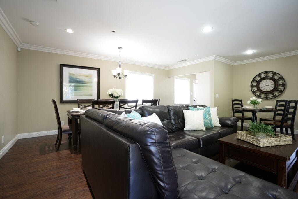 A living room with a sectional sofa, a dining area with a table and chairs, and a wall clock.