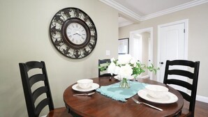 A dining area with a round wooden table, two chairs, a wall-mounted clock, and a vase with flowers. - Classy 4 Bedroom 3 Bath Pool Home 3/4 Mile Walk To Disney (Anaheim)
