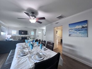 Uma área de refeições com uma mesa posta para seis pessoas, um ventilador de teto e uma pintura de paisagem marítima na parede.