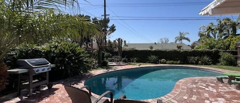 Piscina com um terraço de tijolos, cadeiras de vime e uma churrasqueira.