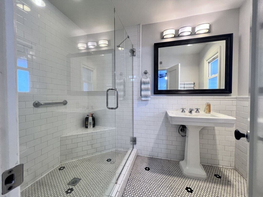A modern bathroom with a glass-enclosed shower, white tiled walls, a white sink, and a large mirror.