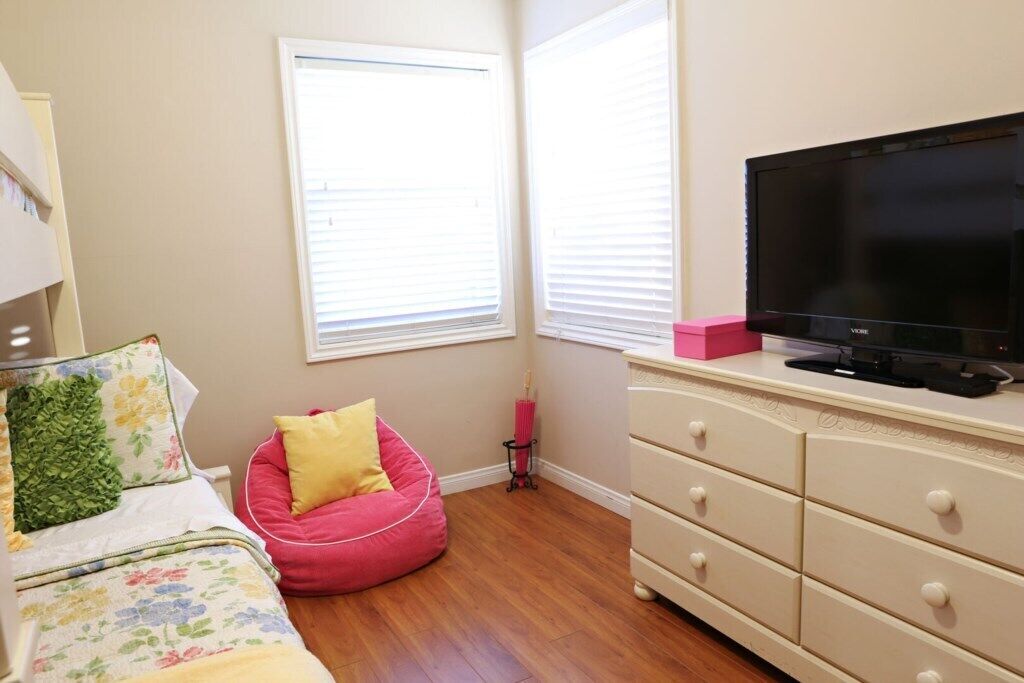A bedroom with a bed, a pink bean bag, a TV on a dresser, and a window with blinds.