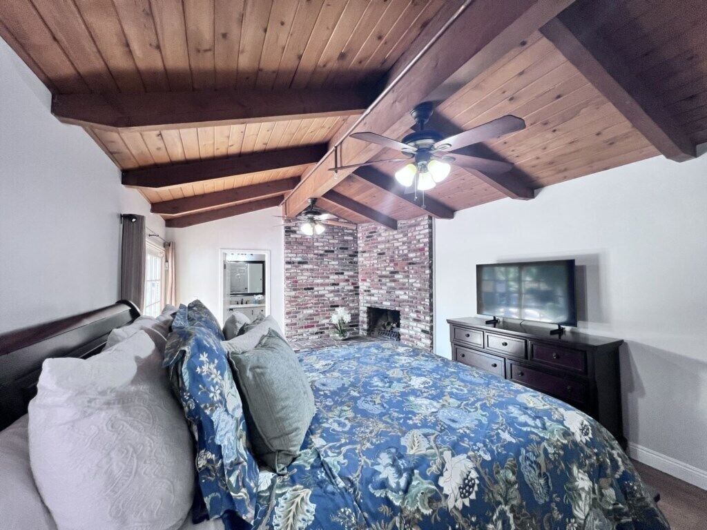 A bedroom with a bed, a TV, a brick wall, and a ceiling fan.