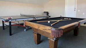 Ein Billardtisch mit Spielzeug und Billardkugeln, ein Tischtennistisch und ein Drucker in einem Raum mit Teppichboden und einer Tür im Hintergrund.