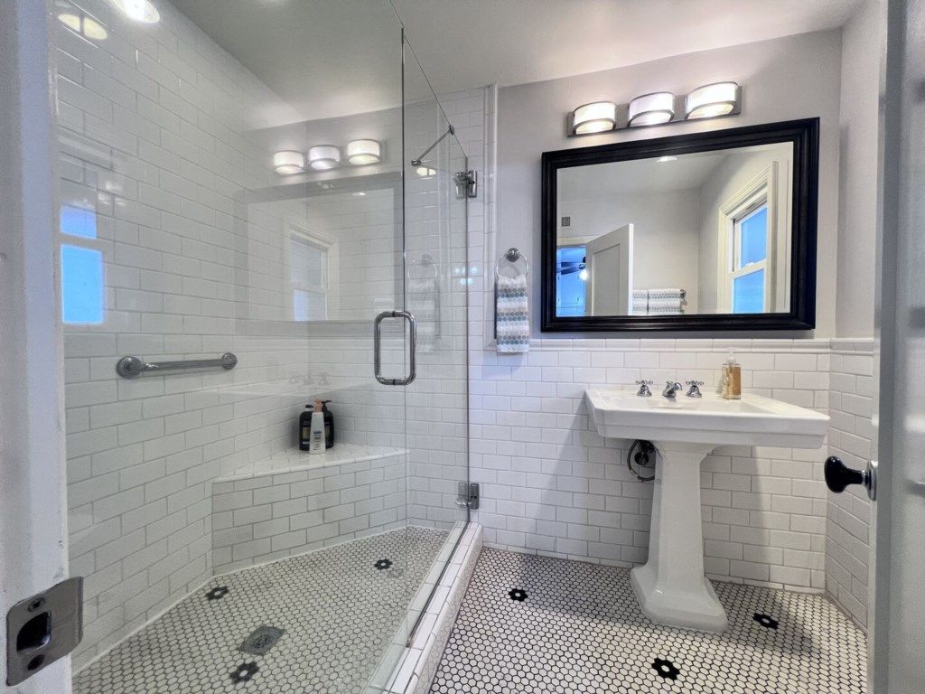 Un baño moderno con ducha de vidrio, paredes con azulejos blancos, un lavamanos blanco y un espejo grande.