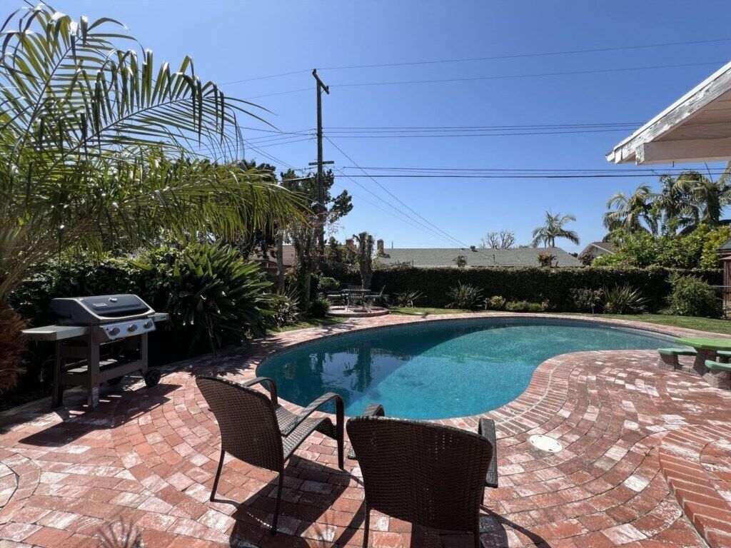 Une piscine avec une terrasse en briques, des chaises en osier et un barbecue.