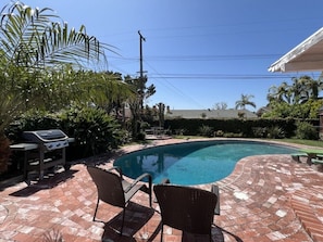 Piscina com um terraço de tijolos, cadeiras de vime e uma churrasqueira.