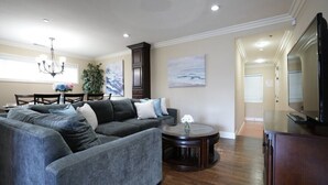A living room with a sectional sofa, a round coffee table, a TV stand, and a dining area with a chandelier. - Comfy Pool Home,1/4 Mile Walk To Disneyland (Anaheim)