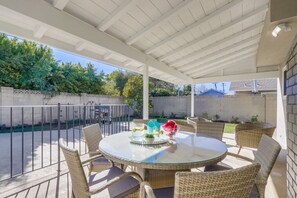 Un patio techado con una mesa redonda de vidrio y sillas de mimbre, rodeado por una cerca blanca y follaje.