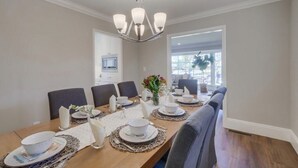 A dining room with a wooden table set for six, surrounded by chairs, a chandelier, and a view of the outdoors.