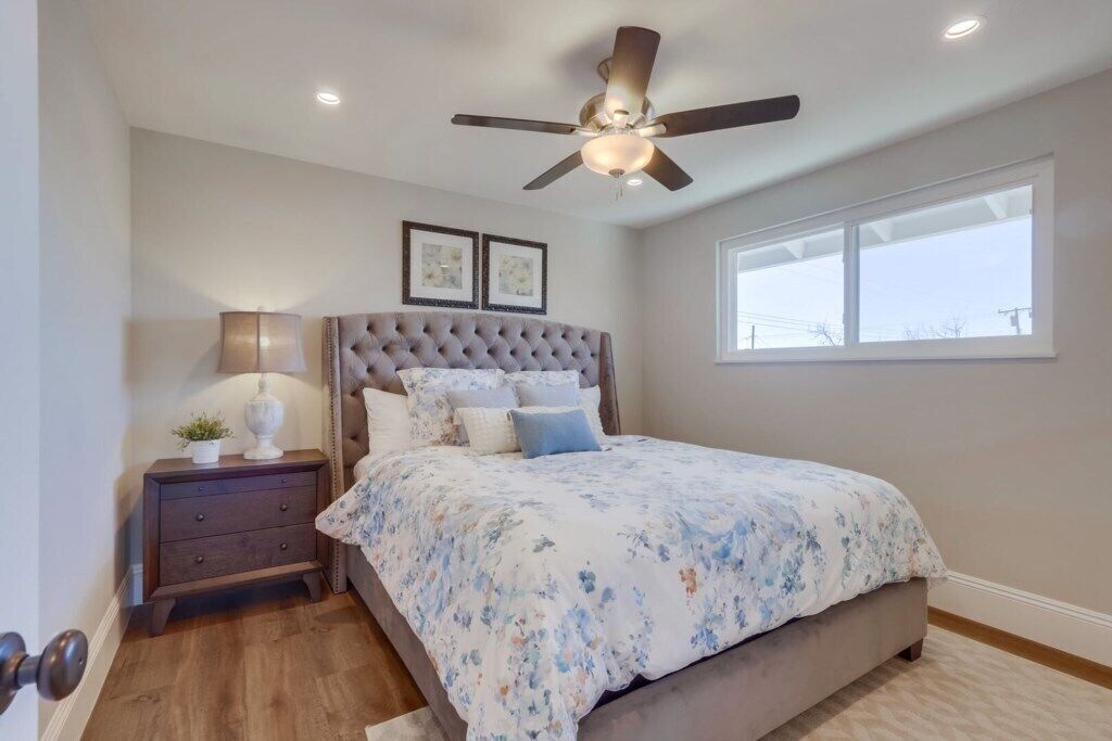 A bedroom with a tufted headboard, a bed with floral bedding, a nightstand with a lamp, a ceiling fan, and a window with a view.