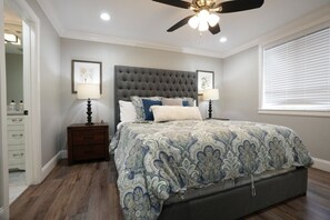 A bedroom with a tufted headboard, two bedside tables with lamps, a bed with a patterned bedspread, a ceiling fan, and a window with blinds.
