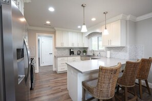 Une cuisine moderne avec des meubles blancs, un îlot central, des chaises en osier et des luminaires suspendus.