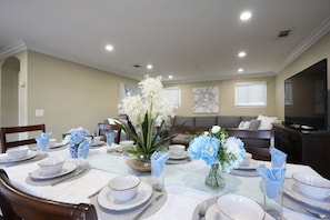 Un coin repas avec une table ronde dressée pour six personnes, entourée de chaises. Des vases décoratifs remplis de fleurs ornent la table.