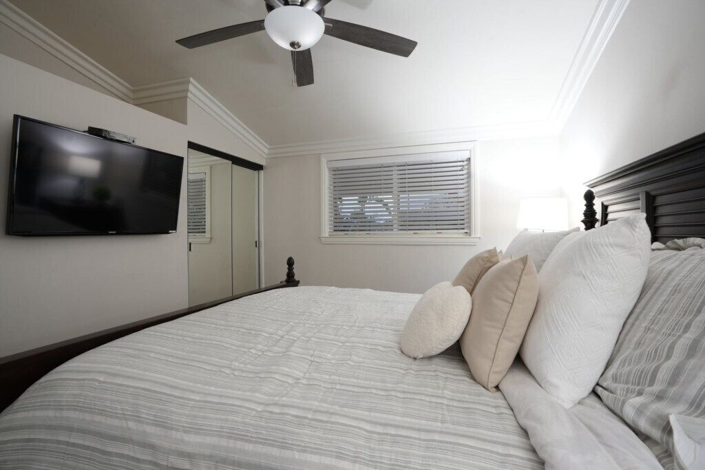Quarto com cama, ventoinha no teto, televisão, janela com persianas e porta.