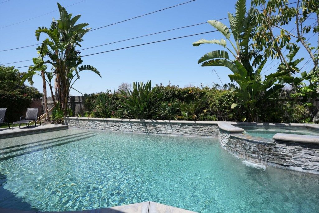 A clear swimming pool with a stone wall, surrounded by lush greenery and a few lounge chairs.