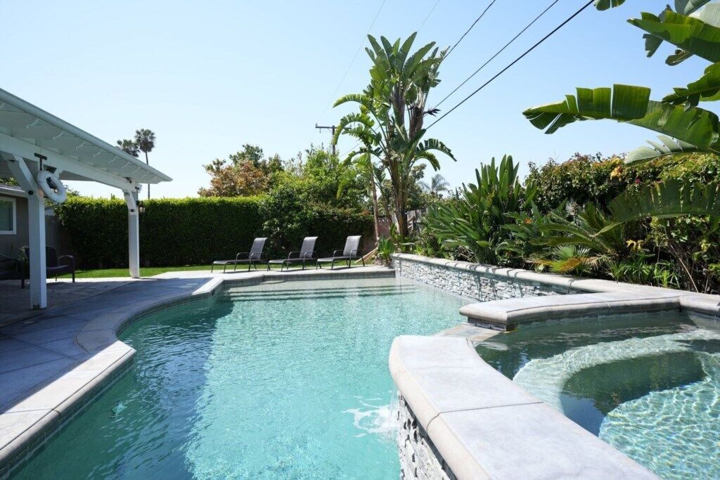A swimming pool with a curved edge, surrounded by a stone deck and a covered patio area with lounge chairs.