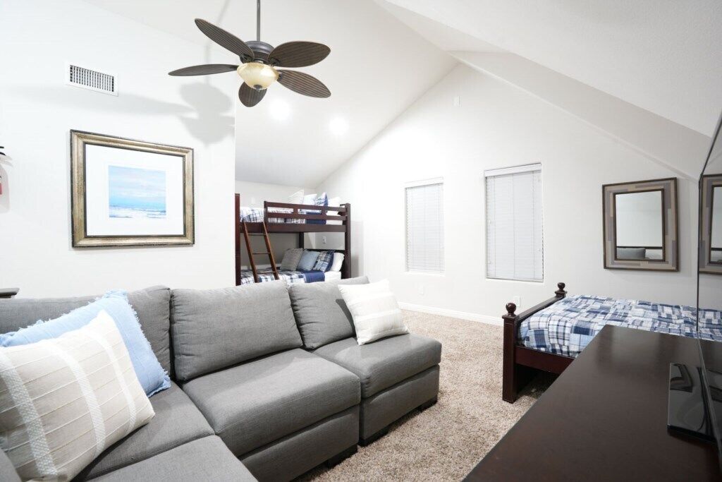 A room with a grey sofa, a bed with a blue and white checkered bedspread, a ceiling fan, and a framed picture on the wall.
