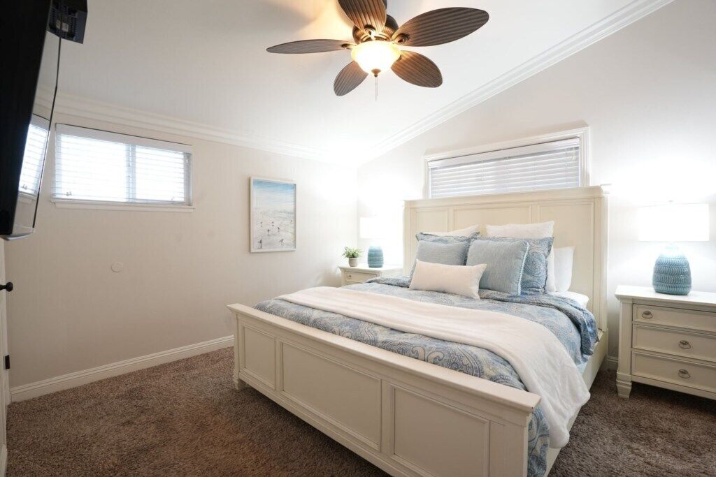 A bedroom with a bed, nightstands, a ceiling fan, a window, and a wall-mounted TV.