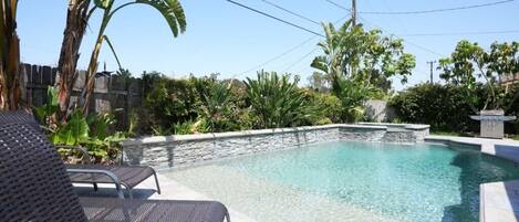 A clear swimming pool with lounge chairs, surrounded by tropical plants and a stone wall.