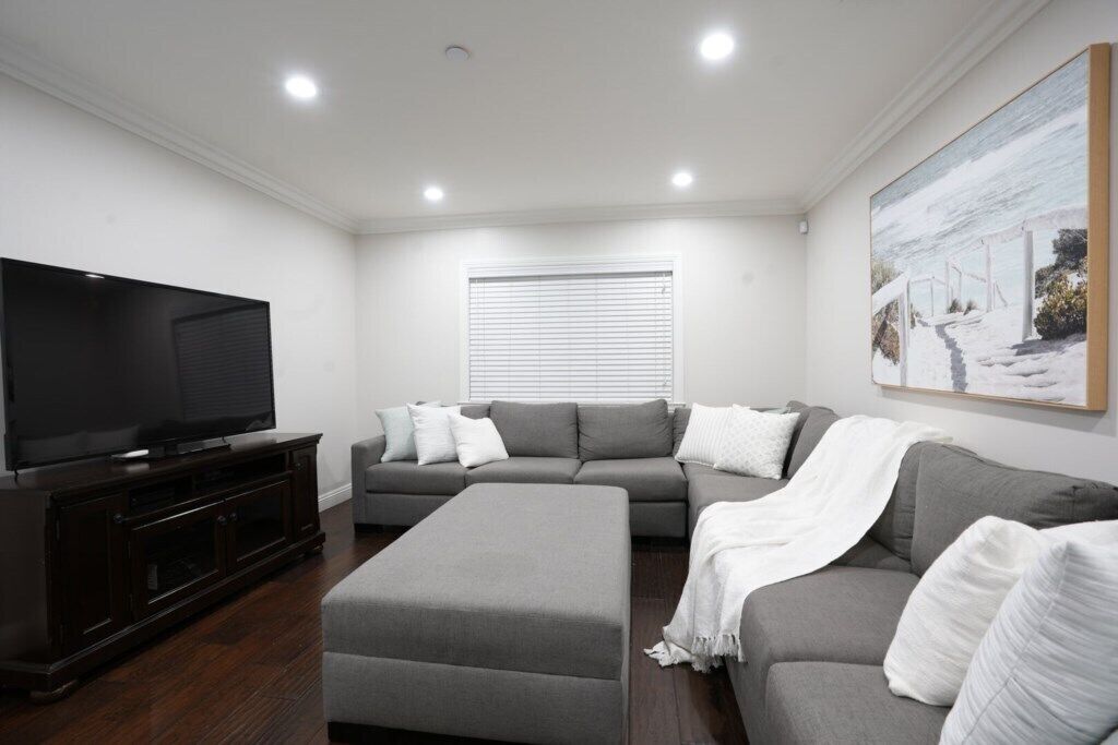 A living room with a flat-screen TV, grey sofas, a white ottoman, and a painting on the wall.