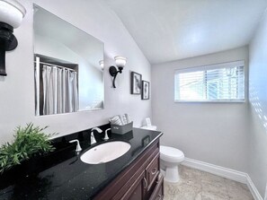 A bathroom with a black countertop sink, a white toilet, a large mirror, and a window with blinds. - Cute 3 Bedroom 2 Bath Home Near Convention Center & Disney (Anaheim)