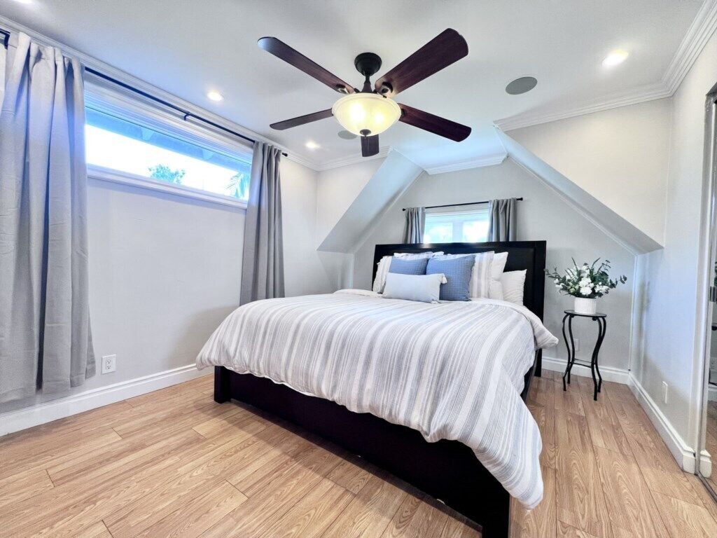 A bedroom with a bed, ceiling fan, window with curtains, and a small table with a plant.
