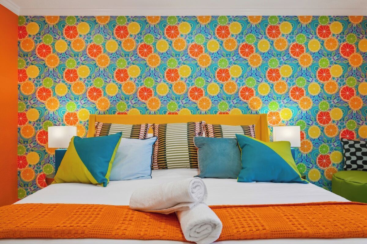 A hotel room with a patterned orange and blue wall, a bed with multiple pillows, and a neatly folded white towel on the bed.