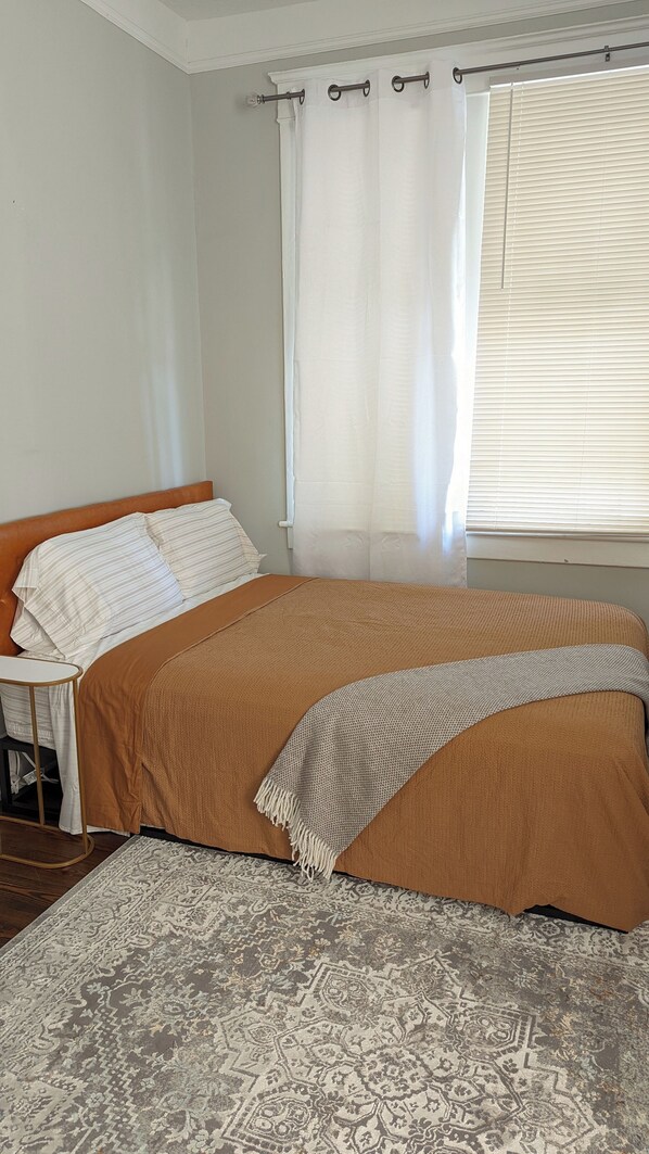 A bedroom with a bed, a nightstand, a window with blinds, and a patterned rug.