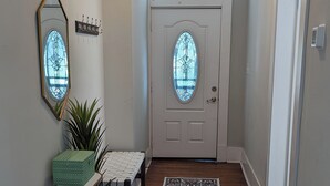 A hallway with a wooden floor, a white door with a glass window, a small table with a plant, and a decorative mirror on the wall. - Mellow ☮ BnB, a NEW 3/1.5 in Victorian District (Savannah)
