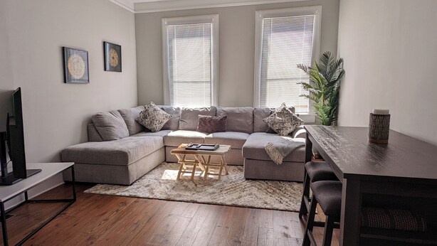 A modern living room with a grey sectional sofa, a wooden coffee table, and a dining area with a wooden table and chairs. - Mellow ☮ BnB, a NEW 3/1.5 in Victorian District (Savannah)